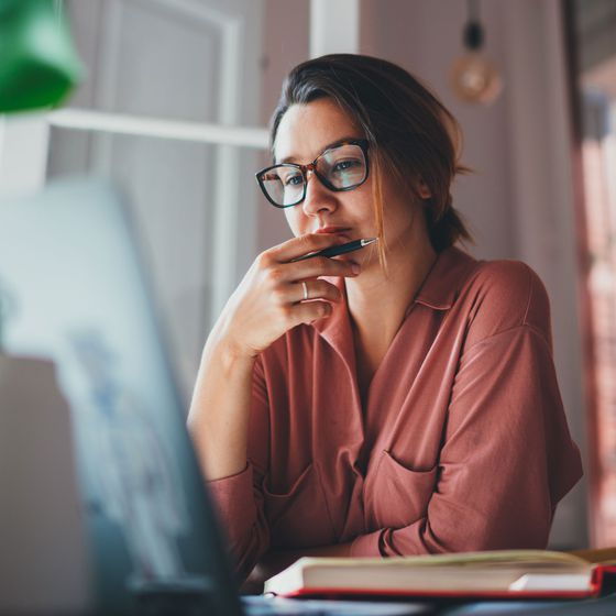 Eine Frau mit rosarotem Hemd sitzt nachdenklich an ihrem Schreibtisch und schaut auf einen Monitor. Vor ihr liegen aufgeschlagene Bücher.